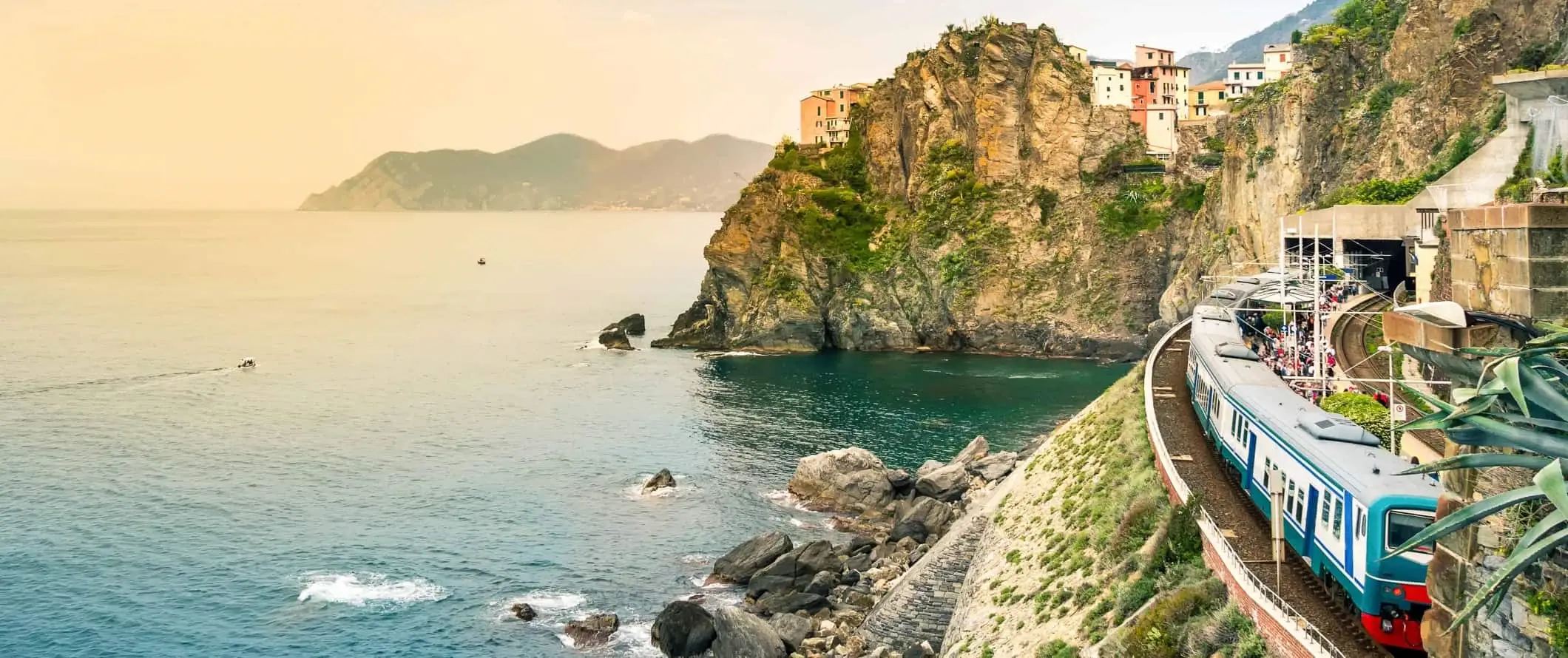 Treno che corre lungo il litorale mediterraneo con scogliere rocciose e villaggi sullo sfondo nelle Cinque Terre, Italia.