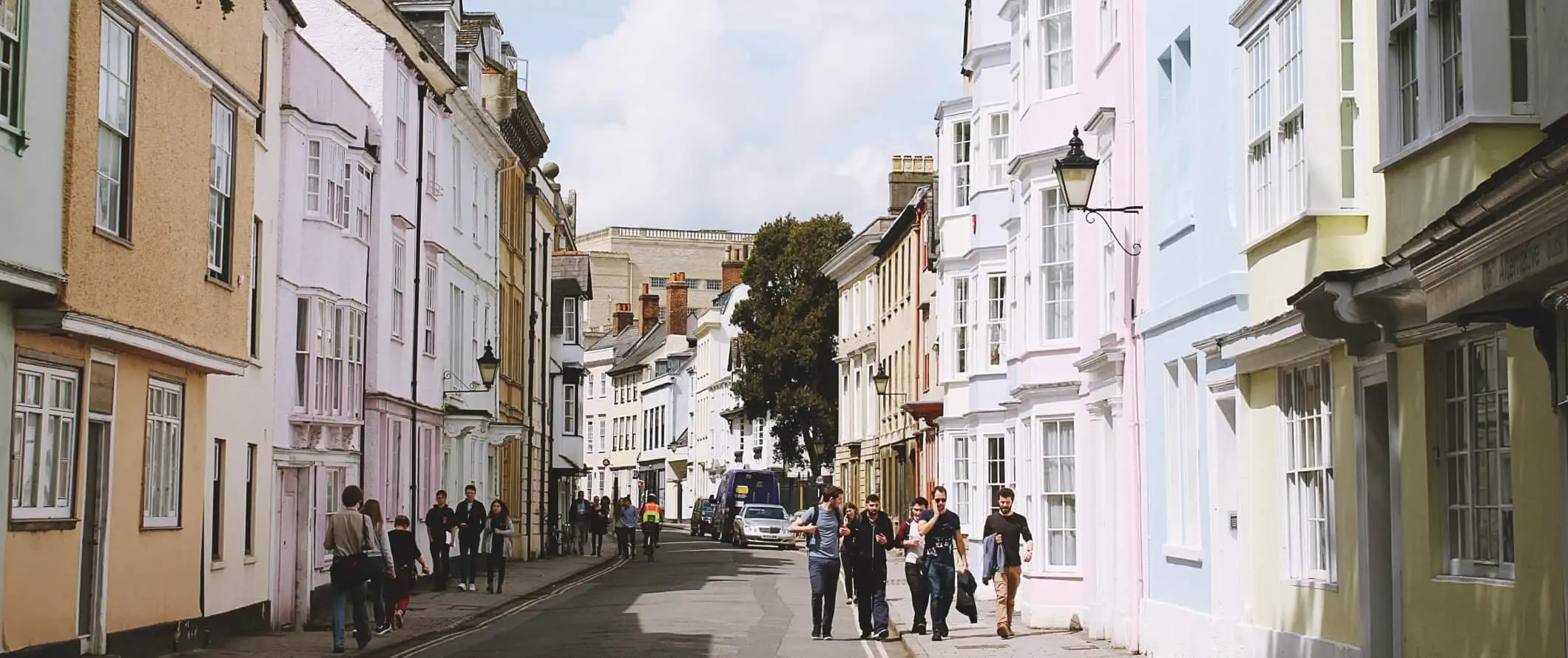 Lidé jdoucí po ulici lemované pastelově zbarvenými městskými domy ve městě Oxford v Anglii