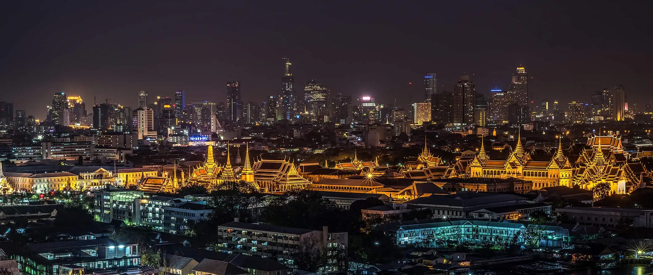 Skyline van Bangkok, Thailand 's nachts, met lage gebouwen op de voorgrond, een tempelcomplex in het midden en moderne wolkenkrabbers op de achtergrond