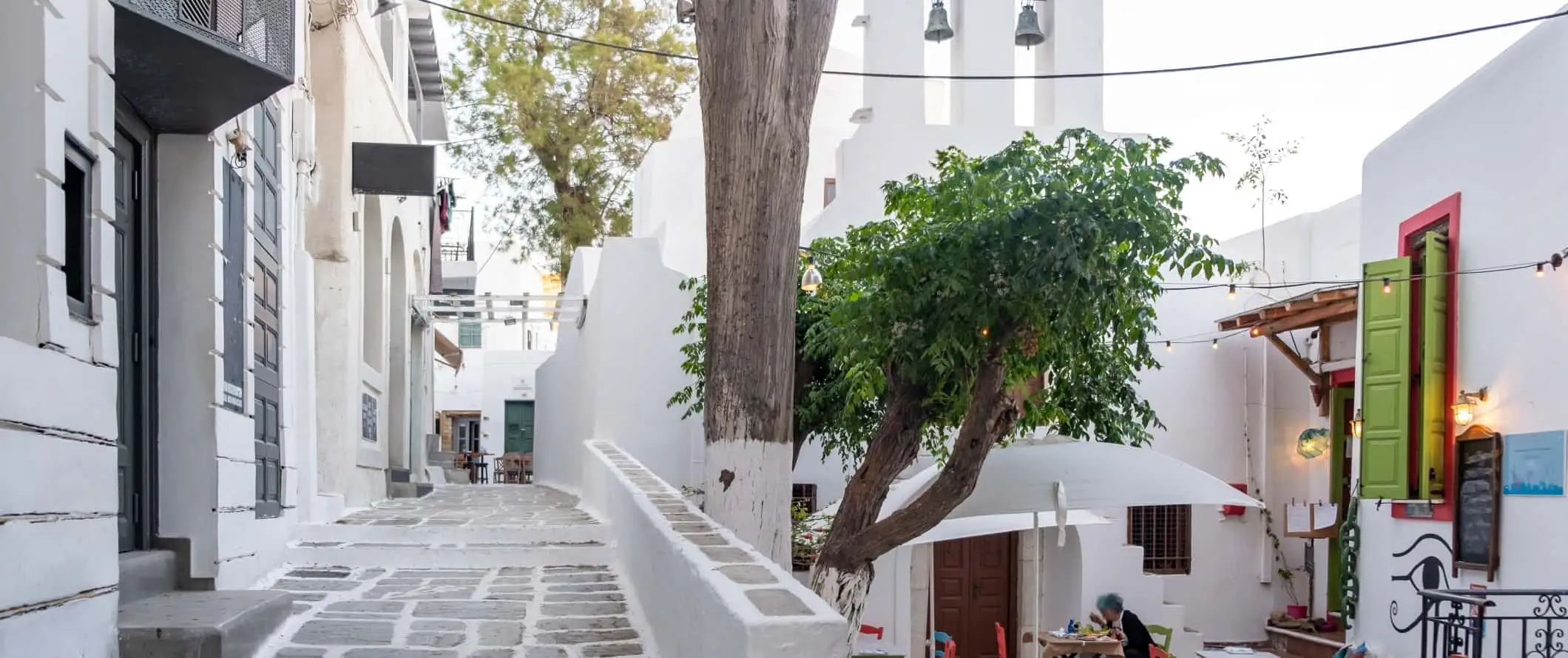 Pemandangan bangunan yang dicuci putih, jalan yang dibarisi batu bendera dan tempat duduk di luar di kafe dengan meja dan kerusi berwarna terang di Ios, Greece