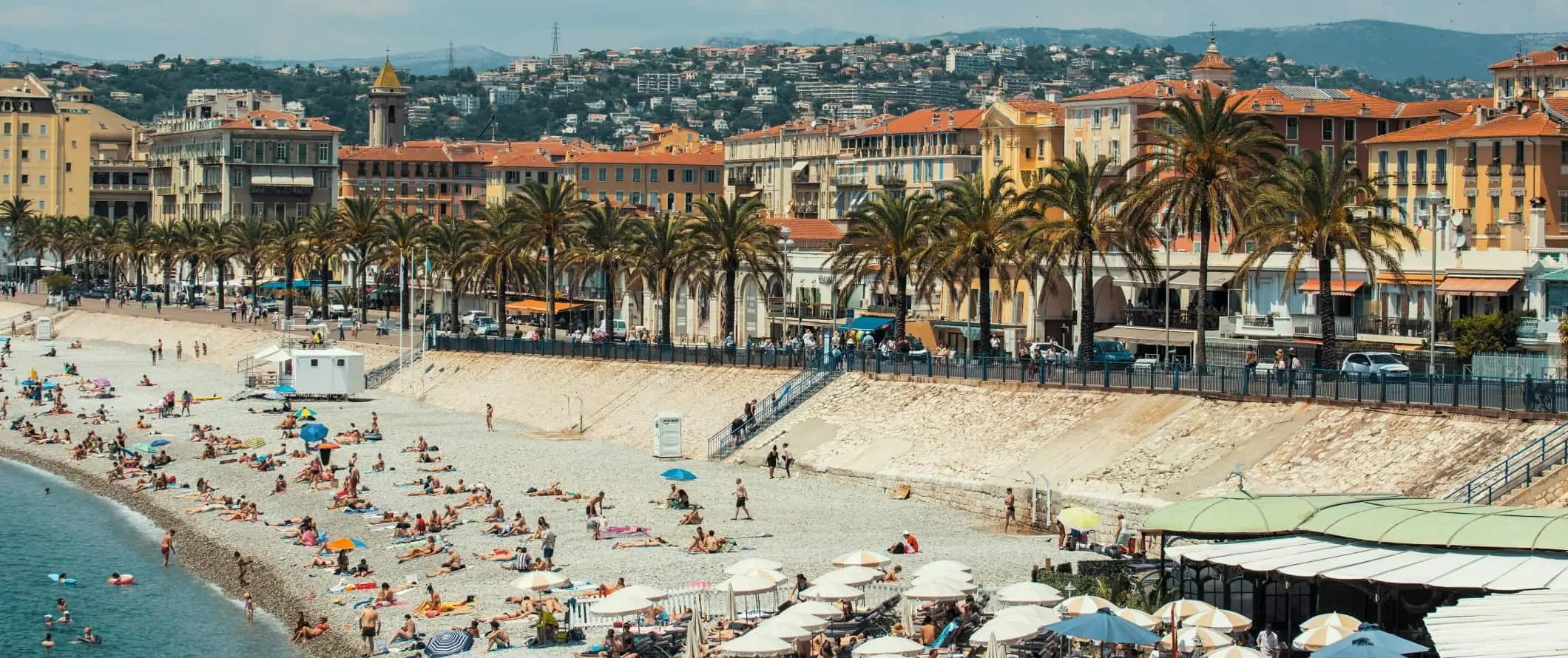 Lidé ležící na pláži před promenádou lemovanou palmami a v pozadí se tyčí město Nice, Francie