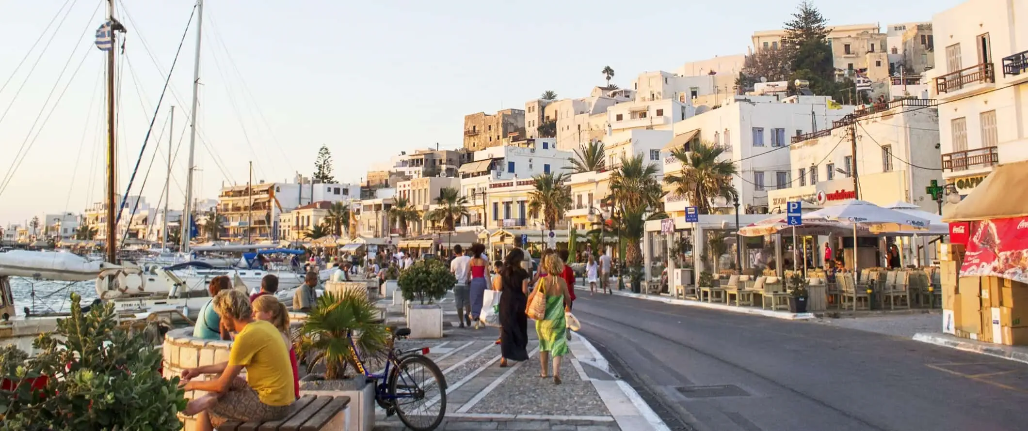 Folk som sitter på benker og går langs havnefronten fylt med seilbåter, og Chora gamleby med sine hvitkalkede bygninger i bakgrunnen på øya Naxos i Hellas.