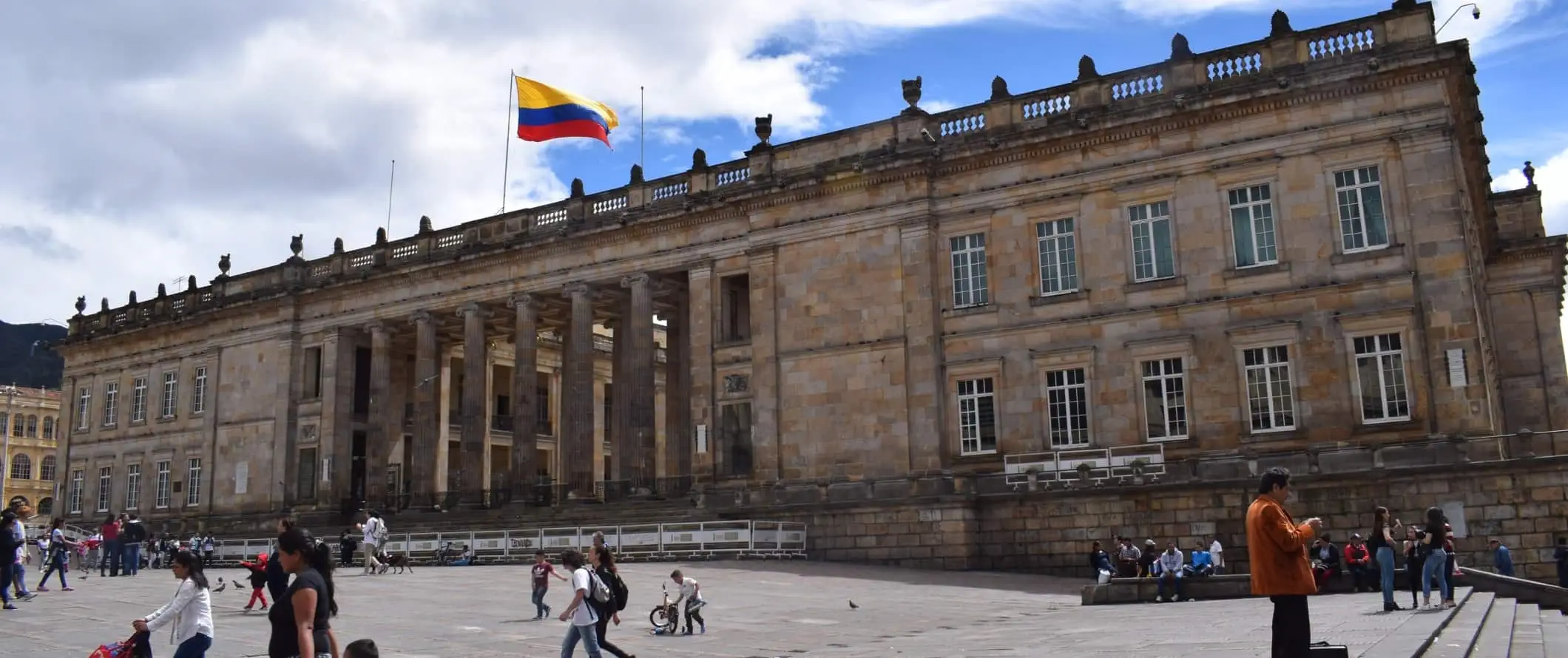 Mensen lopen rond voor een groot neoklassiek gebouw met veel pilaren en de Colombiaanse vlag die vanaf het dak wappert in Bogota, Colombia