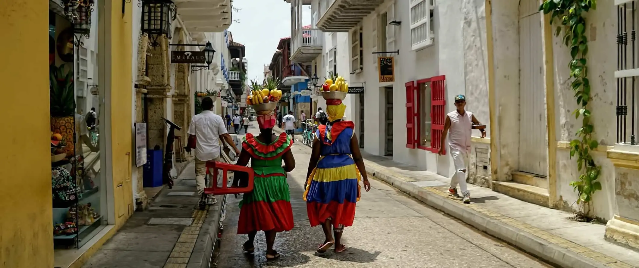 To kvinner i lyse, fargerike kjoler går nedover en gate med fruktkurver på hodet i Cartagena, Colombia