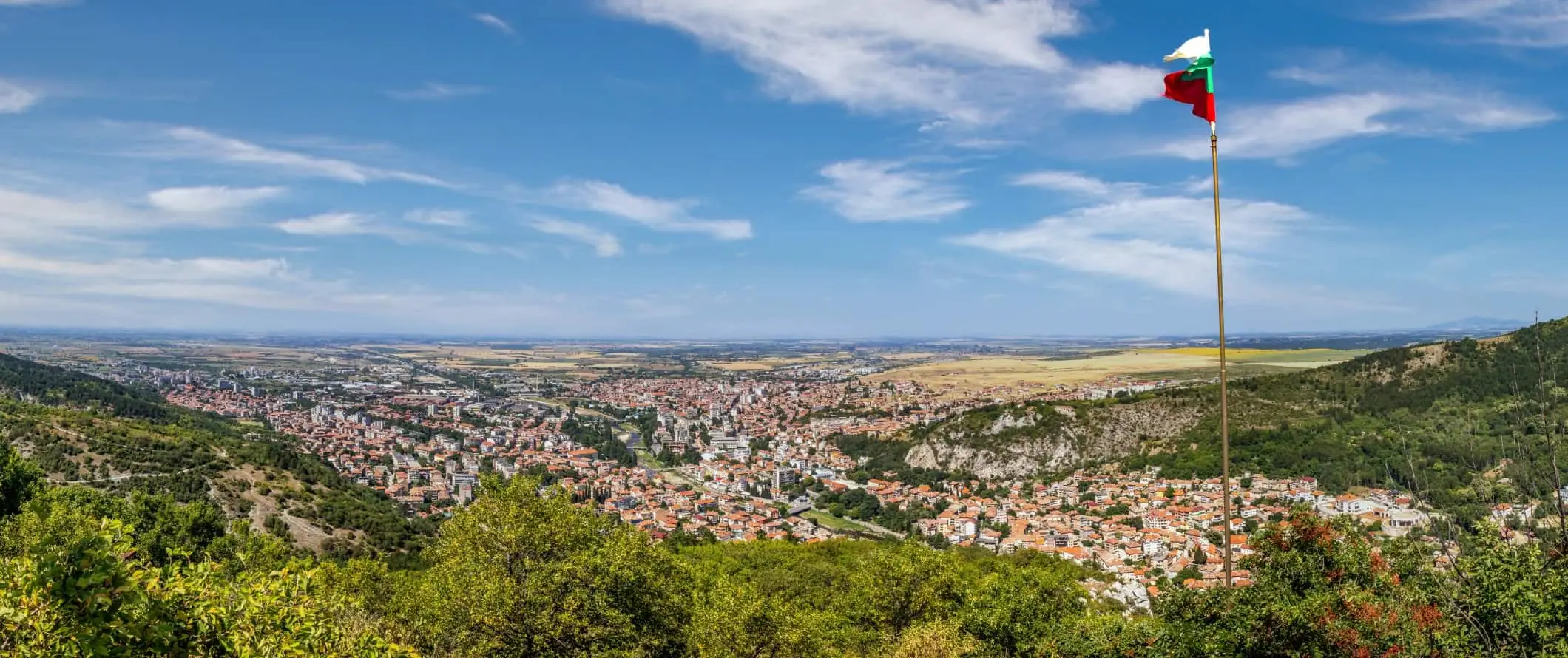 Panoramautsikt över de röda terrakottataken i den historiska staden Asenovgrad mitt bland böljande kullar i Bulgarien, med den bulgariska flaggan i förgrunden