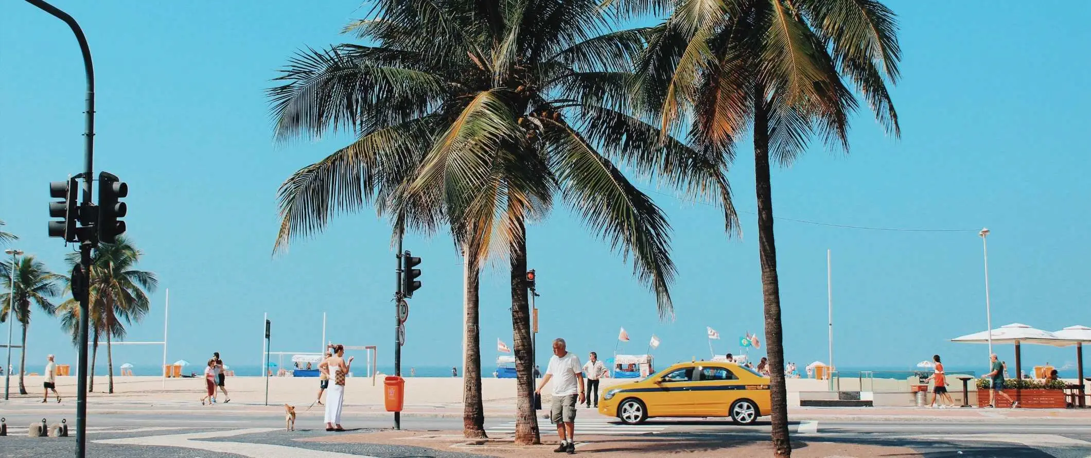 Menschen, die mit einem gelben Taxi am Strand von Rio de Janeiro, Brasilien, eine von Palmen gesäumte Straße entlanglaufen