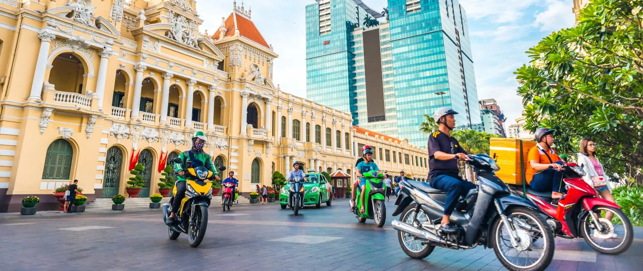 Vietējie iedzīvotāji brauc ar motorolleriem un mopēdiem uz rosīgas ielas Hošiminā, Vjetnamā pie rātsnama