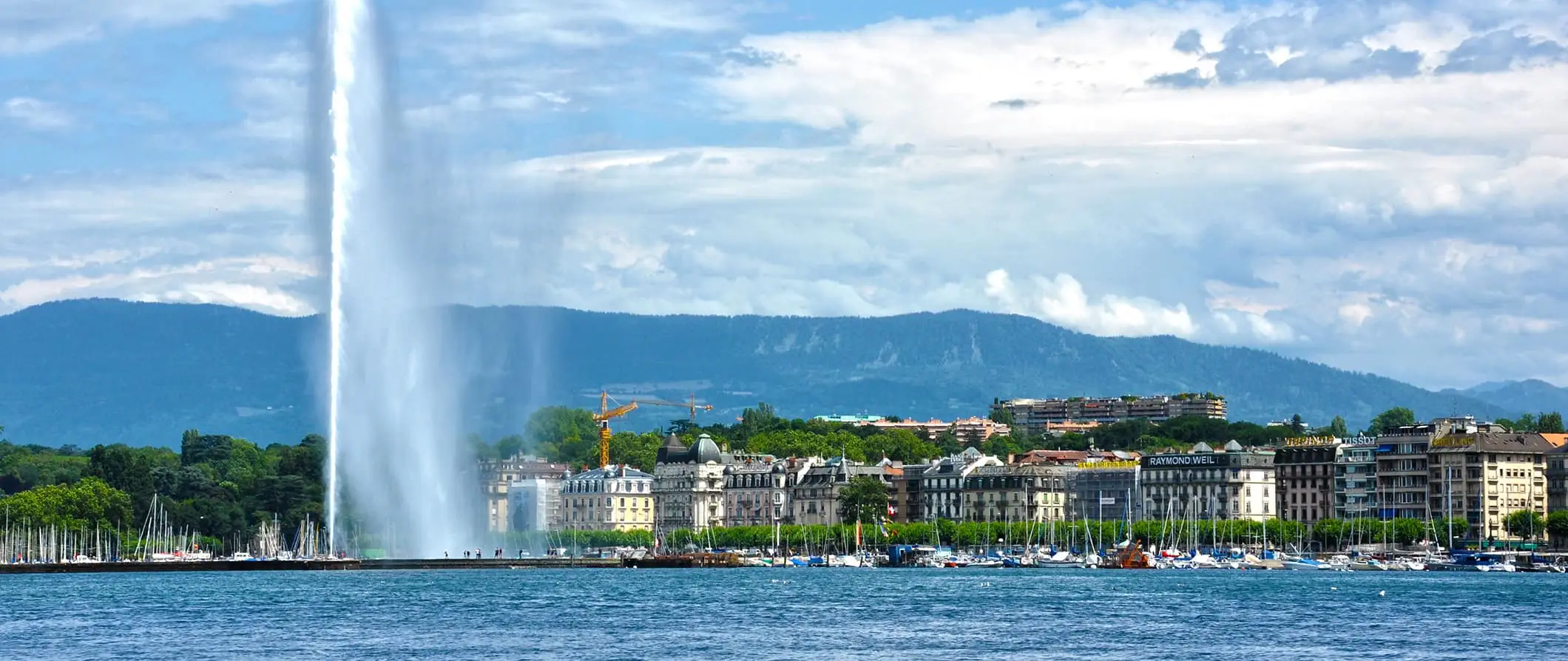 Blick auf den Jet-Brunnen und die Uferpromenade in Genf, Schweiz, an einem hellen und sonnigen Sommertag mit Bergen in der Ferne