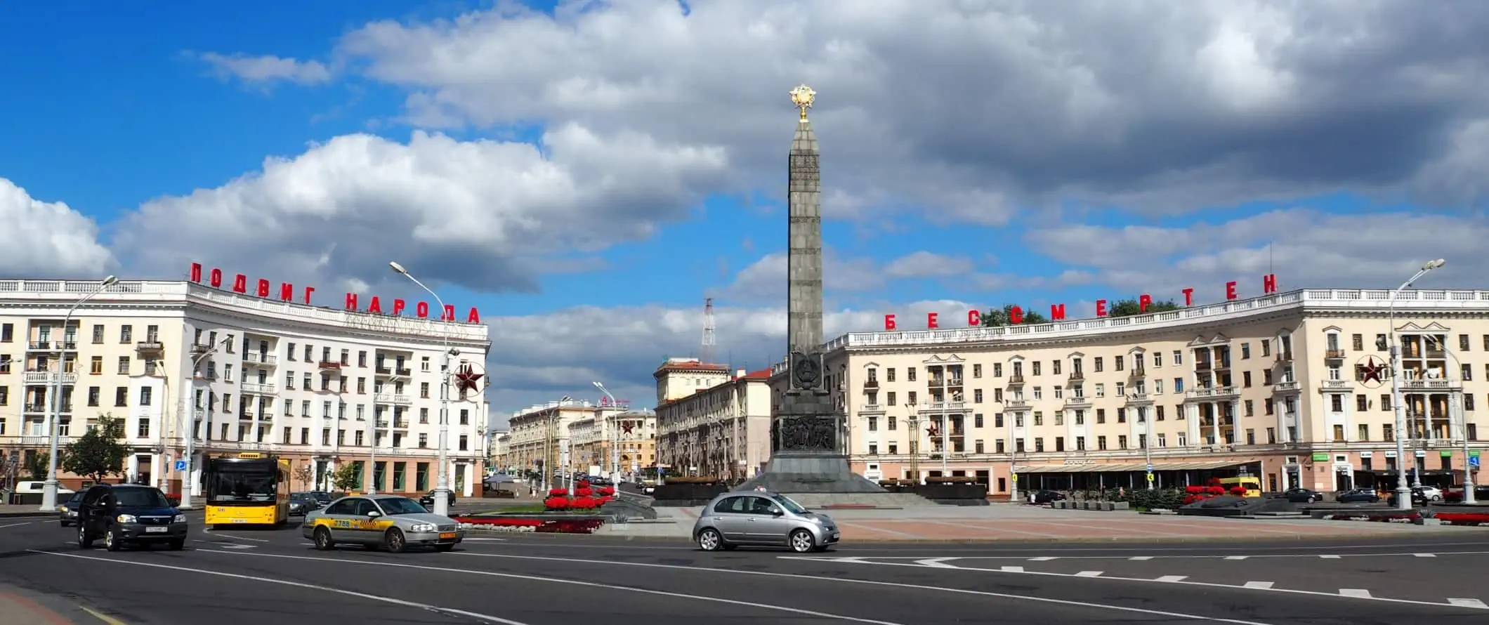 Autók körbejárnak egy nagy körforgalmat a Győzelem tér előtt Minszkben, Fehéroroszországban