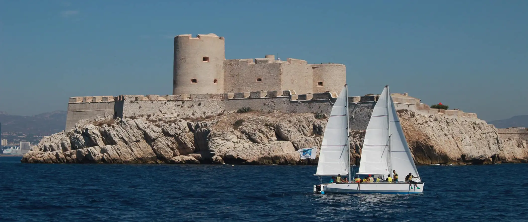 Un pequeño velero lleno de pasajeros pasando frente al castillo de If, ​​una fortaleza frente a la costa de Marsella.