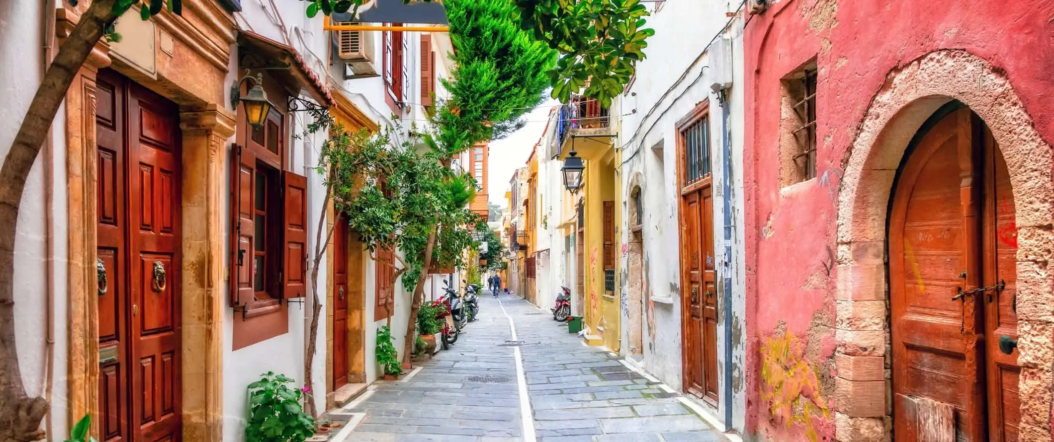 Jalan yang dibarisi batu bendera diapit oleh rumah berwarna terang dengan pintu kayu di pulau Crete di Greece.