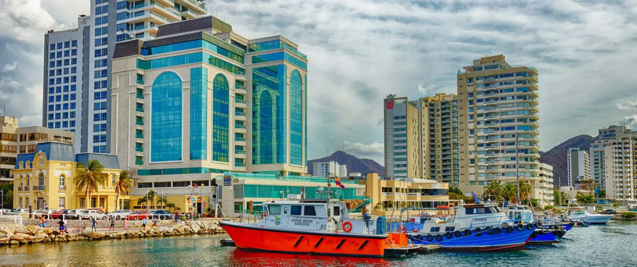 Bunte Boote im Hafen mit Wolkenkratzern im Hintergrund in der Stadt Santa Marta, Kolumbien