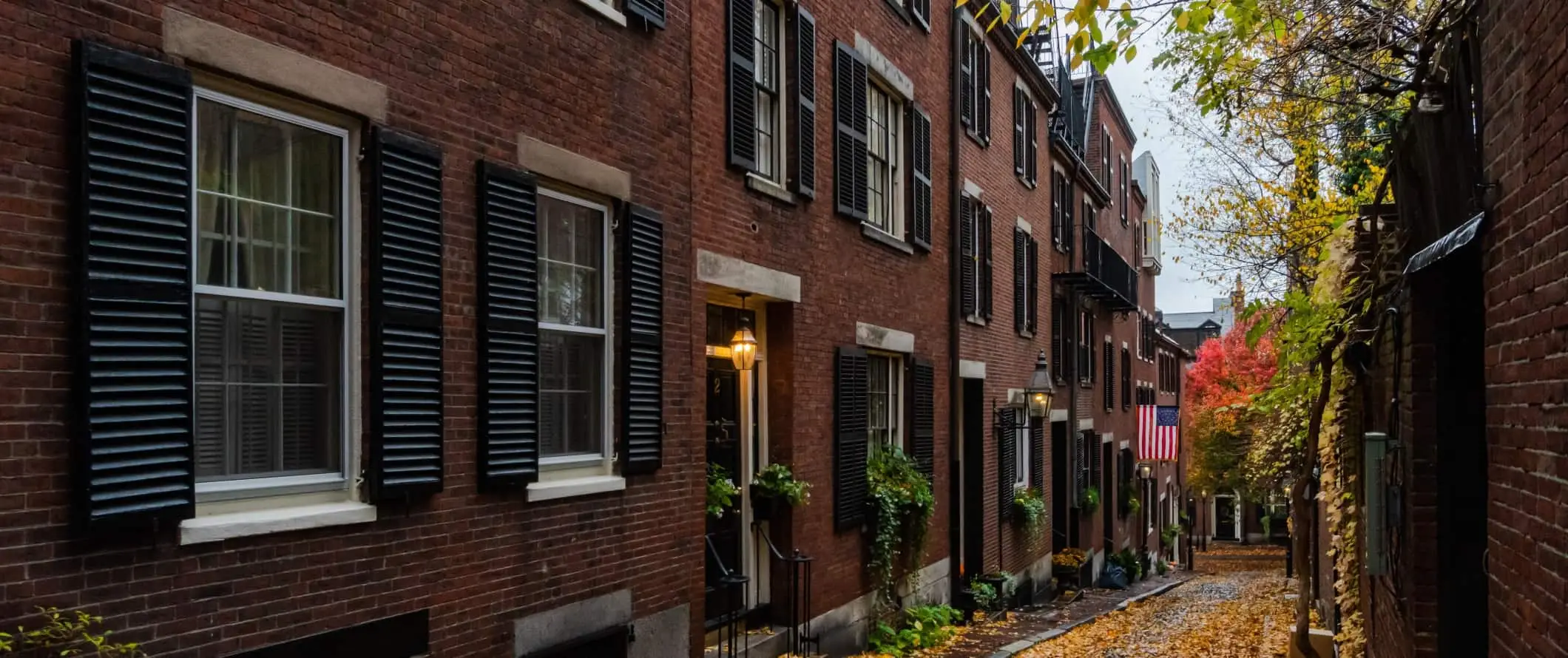 Historiske murstenshuse med sorte skodder langs en gyde med orange blade på jorden i Boston, Massachusetts.